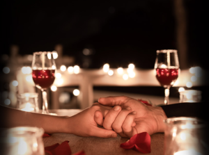 imagem mostra duas mãos juntas e duas taças de vinho ao fundo