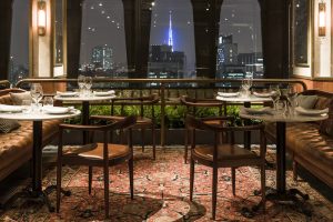 imagem mostra um restaurante com mesas e cadeiras de madeira, com carpete vermelho e visão noturna para a cidade