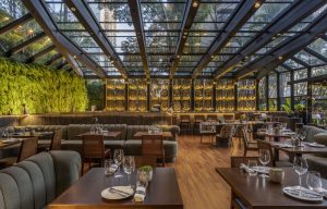 imagem mostra ambiente de restaurante com mesa, parede revestida de plantas e teto de vidro