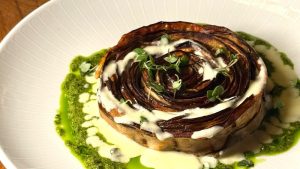 Prato Fiori de Melanzane com berinjela assada em formato de flor, servido com molho cremoso e azeite de ervas, finalizado com brotos verdes