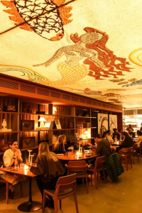 Restaurante com iluminação aconchegante e mesas ocupadas por clientes. No teto, uma grande ilustração artística de uma sereia em tons terrosos se destaca sobre o ambiente, que combina estantes decorativas, móveis de madeira e atmosfera elegante.