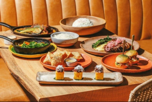 Mesa de madeira com diferentes pratos do novo menu do Terraço Jardins, incluindo petiscos, sanduíches, carnes, acompanhamentos e sobremesas servidos em louças coloridas.