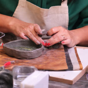 Pessoa trabalha manualmente na modelagem de uma peça de cerâmica sobre uma mesa de madeira. As mãos moldam a argila enquanto uma pequena peça em formato de recipiente toma forma. Ao redor, estão materiais utilizados na oficina, como ferramentas e recipientes de apoio. A imagem representa a experiência Ateliê Cerâmica & Vinho, que une arte, criatividade e bem-estar em um ambiente descontraído.