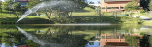 Vista de um lago cercado por áreas verdes no Almenat by Hilton, em Embu das Artes. Um sistema de irrigação lança um arco de água sobre o lago, enquanto árvores e edificações do hotel aparecem ao fundo refletidas na superfície da água.