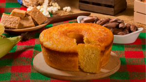 Mesa com comidas típicas de festa junina, incluindo bolo de milho, doces, pipoca e pinhões, dispostos sobre toalha xadrez colorida em ambientação temática de São João.