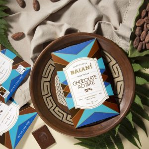Embalagem de chocolate da Baianí Chocolates posicionada sobre uma bandeja de madeira, cercada por amêndoas de cacau, folhas e elementos decorativos. A imagem destaca uma barra de chocolate produzida com cacau cultivado no Vale Potumuju, na Bahia, evidenciando a proposta bean to bar da marca.
