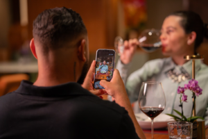 Pessoa fotografa o companheiro durante um jantar em restaurante. Sobre a mesa, taças de vinho, flores e elementos de decoração criam uma atmosfera romântica e acolhedora.