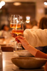 A imagem mostra duas pessoas brindando com taças de vinho rosé ou espumante rosé em um ambiente interno aconchegante. A iluminação é quente, com efeito de desfoque ao fundo (bokeh), criando uma atmosfera elegante e intimista. As mãos das pessoas aparecem em destaque segurando as taças, enquanto o restante do restaurante ou salão permanece suavemente desfocado.